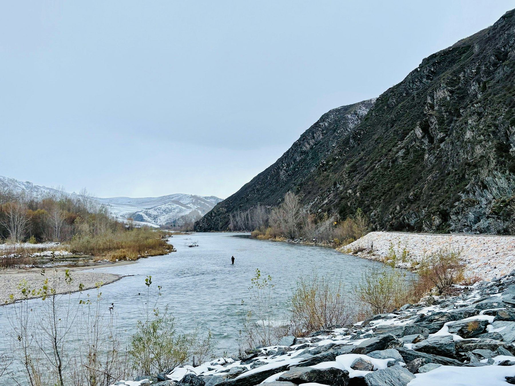 A fly fisherman casting in a river below snow-covered peaks
